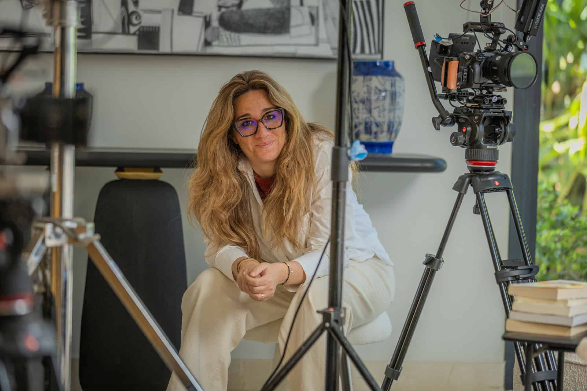 Woman with long blonde hair and glasses sitting in a modern studio surrounded by professional filming equipment.