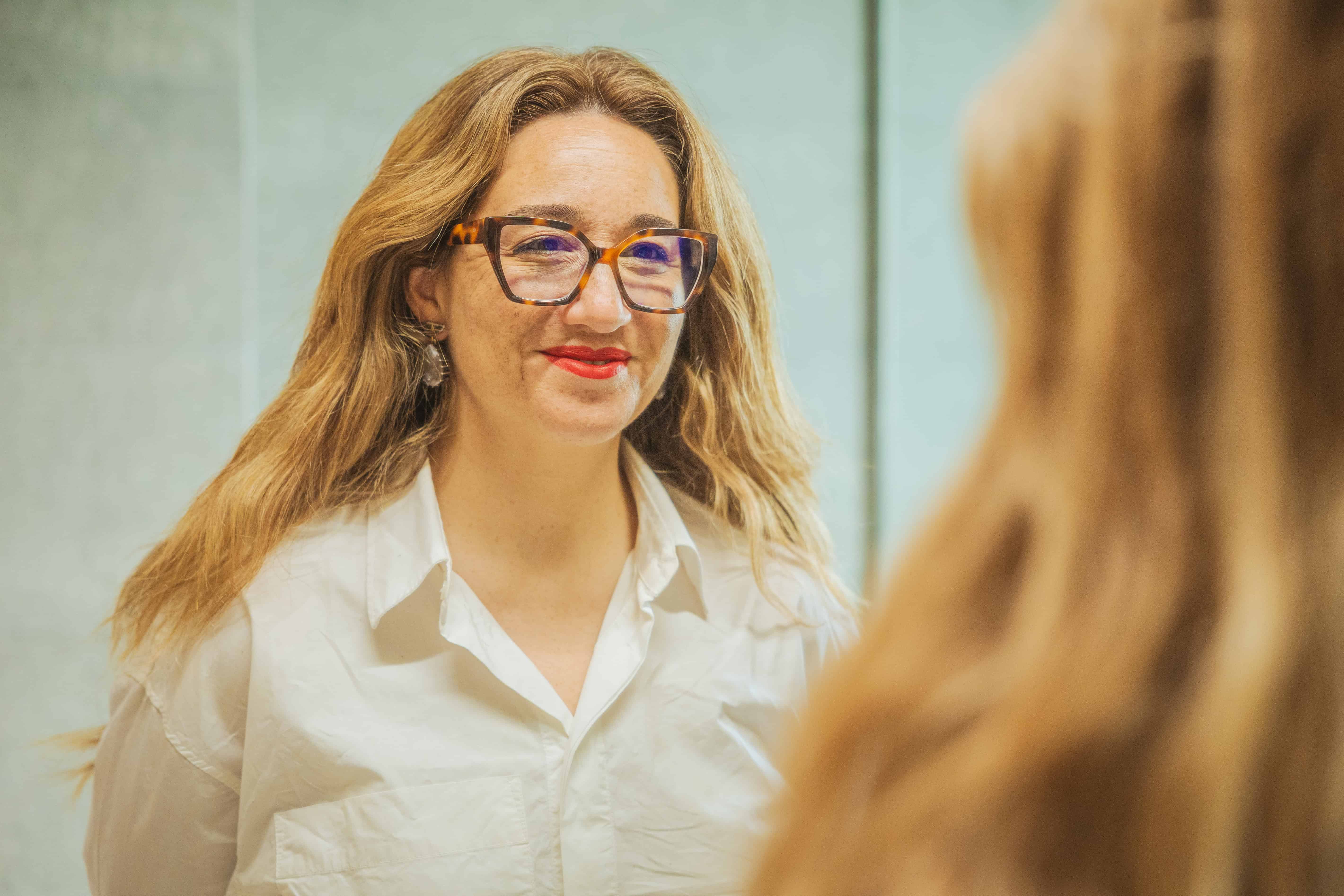 Professional portrait of a woman with glasses and blonde hair, smiling in a business setting, representing leadership and expertise.