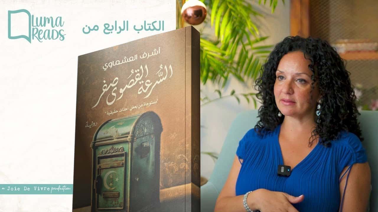 A woman with curly black hair wearing a blue dress seated indoors next to a book titled "الكتاب الرابع من أشرف العشماوي".